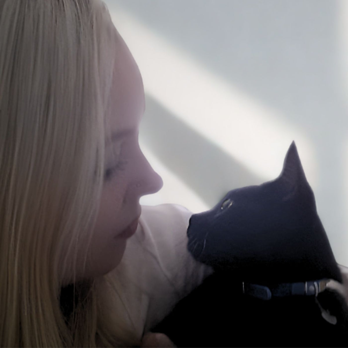 A close-up lifestyle shot of a blonde woman in profile hugging a black cat wearing a blue collar, set against a bright, sunlit white wall.