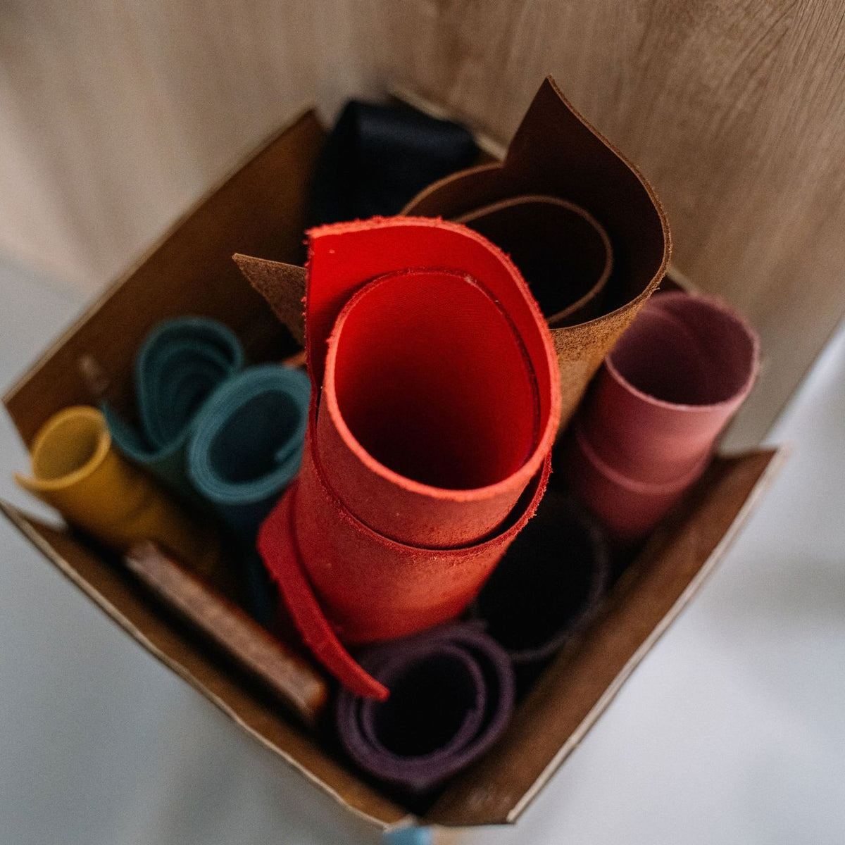 A cardboard box filled with rolled leather sheets in multiple colors, including red, brown, teal, pink, yellow, and purple, photographed from above.
