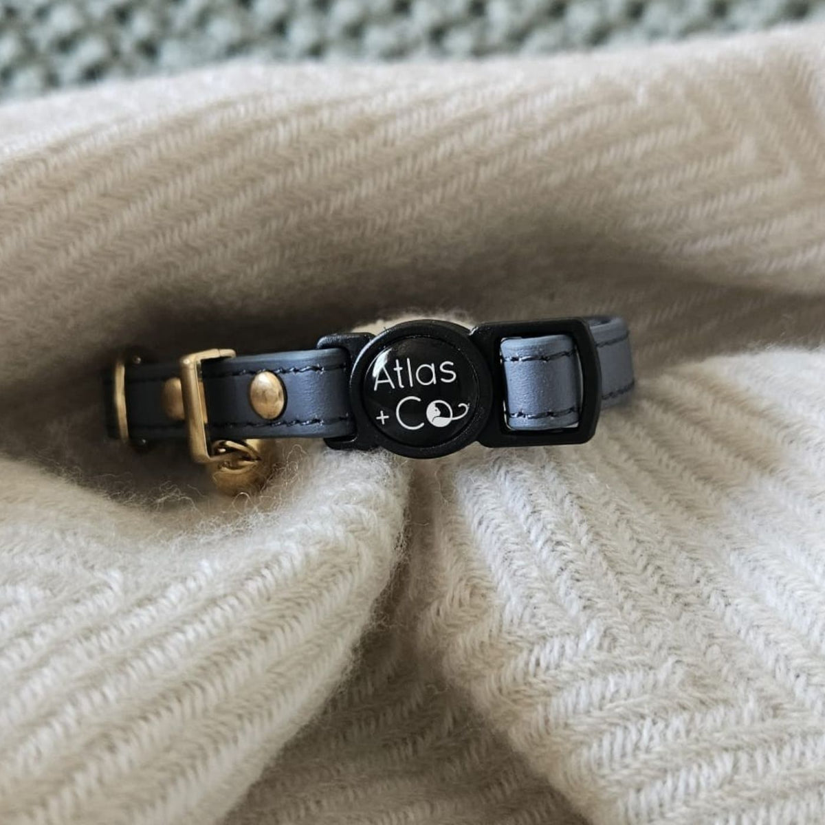 A close-up of a blue leather cat collar with a gold-tone buckle and a central round black tag engraved with 'Atlas + Co', resting on a cream-colored ribbed knit blanket.