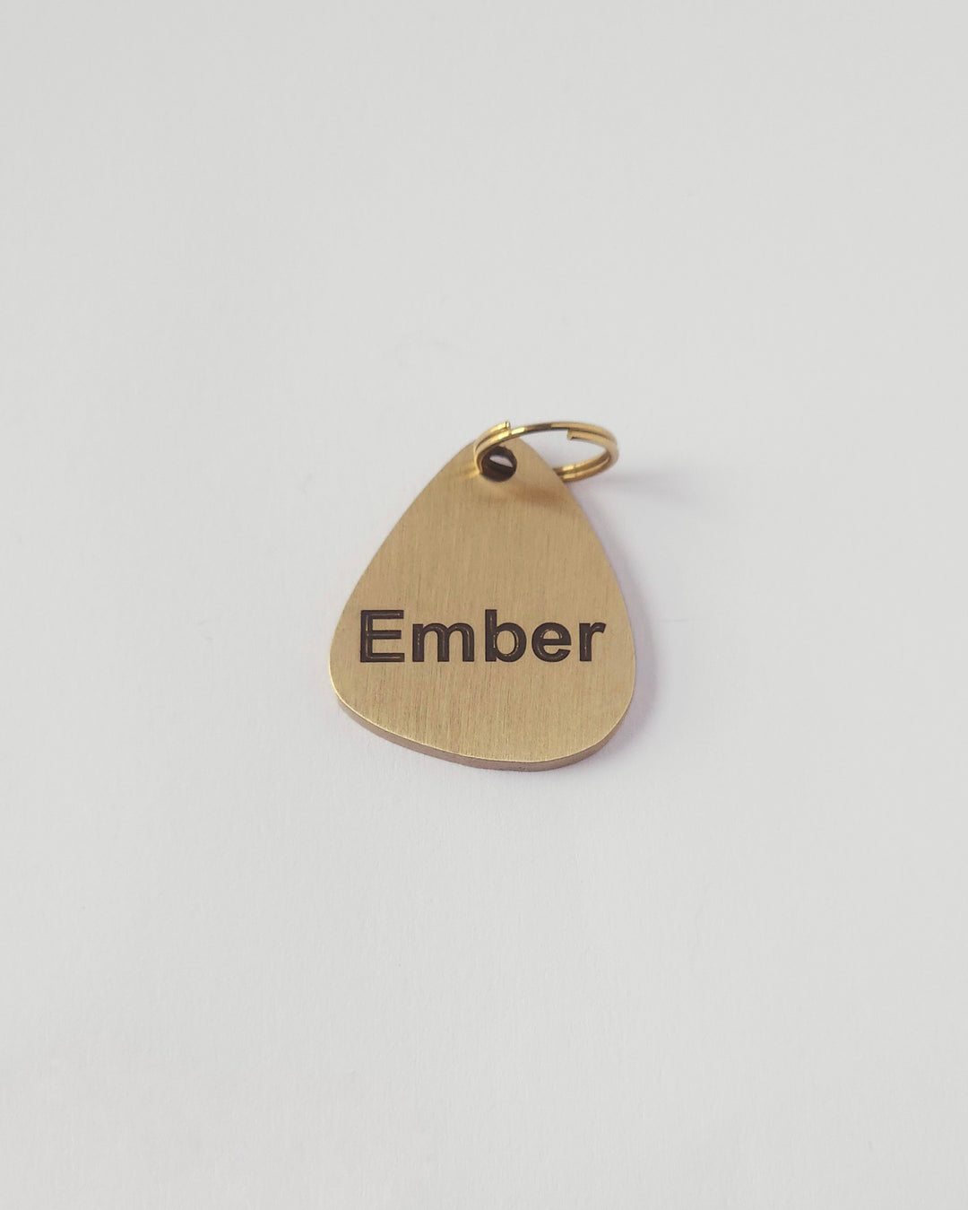 A teardrop-shaped gold metal pendant engraved with the name 'Ember' and featuring a small hanging loop at the top, shown on a plain light gray background. #shape_pebble