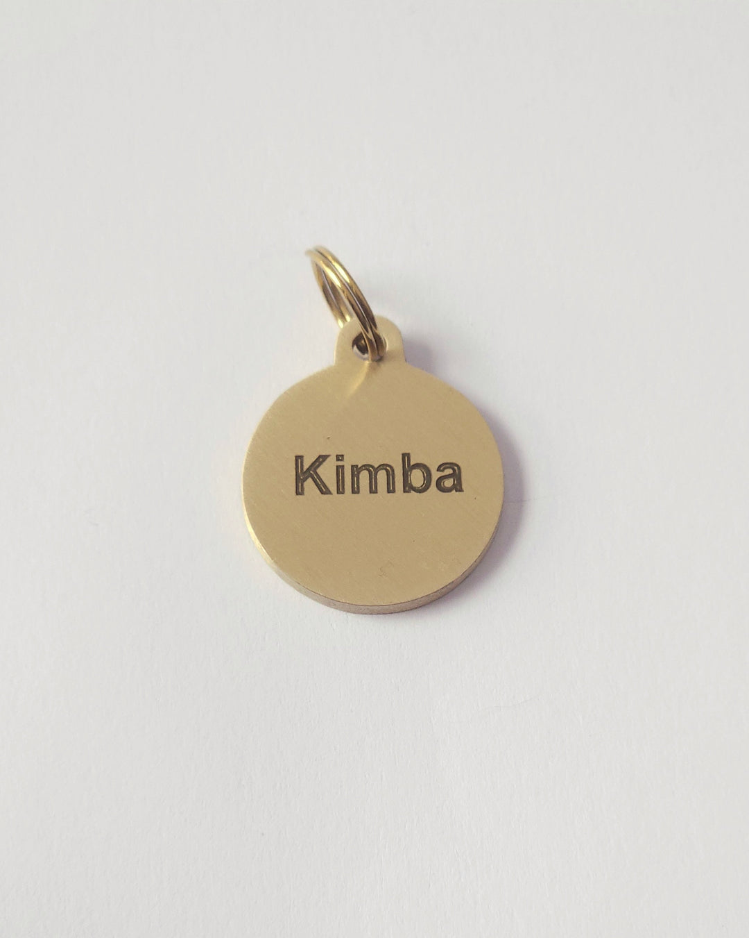 A round brass metal charm pendant engraved with the name 'Kimba' in black lowercase letters, shown on a plain white background. #shape_round