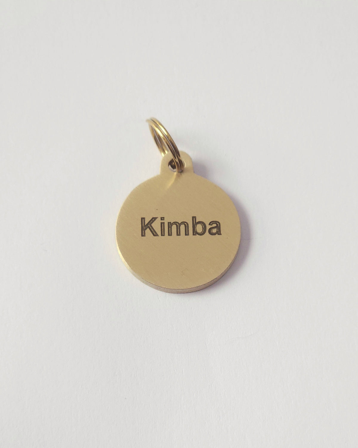 A round brass metal charm pendant engraved with the name 'Kimba' in black lowercase letters, shown on a plain white background. #shape_round