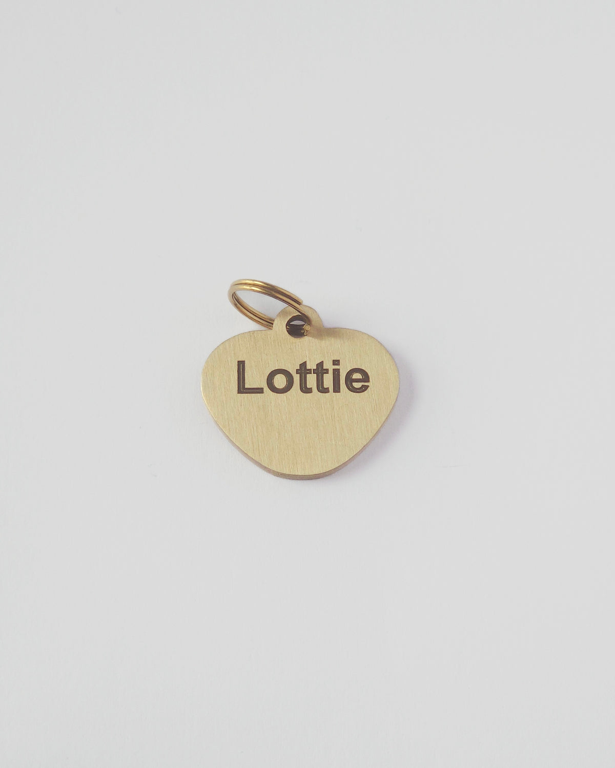 A small heart-shaped brass pet tag engraved with the name 'Lottie' and attached to a matching split ring, shown on a clean light gray background. #shape_roundedheart